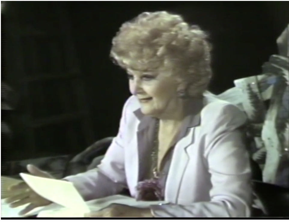An older video still of a smiling woman with voluminous curly blonde hair wearing a light pink blazer over a mauve blouse, seated at a desk and looking at a piece of paper in her hands.
