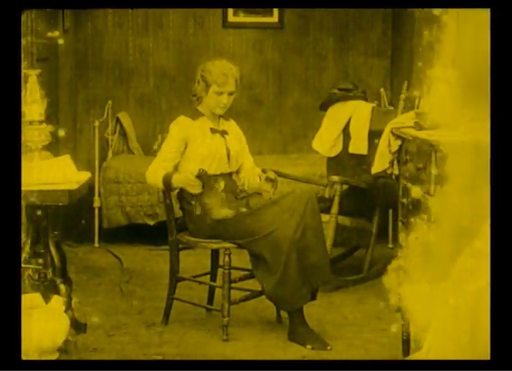 A yellow-tinted still from a silent film showing a young woman with short hair sitting on a wooden chair in a modest bedroom, holding a dark object, possibly a shoe, in her lap.