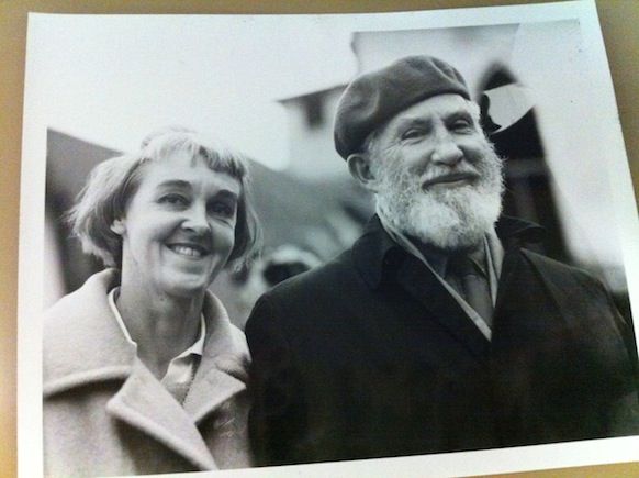 A black and white portrait photograph of a smiling older couple outdoors. The woman on the left has short light hair with bangs and wears a light-colored coat. The man on the right has a full white beard, is smiling, and wears a dark beret, a dark coat, and a tie.