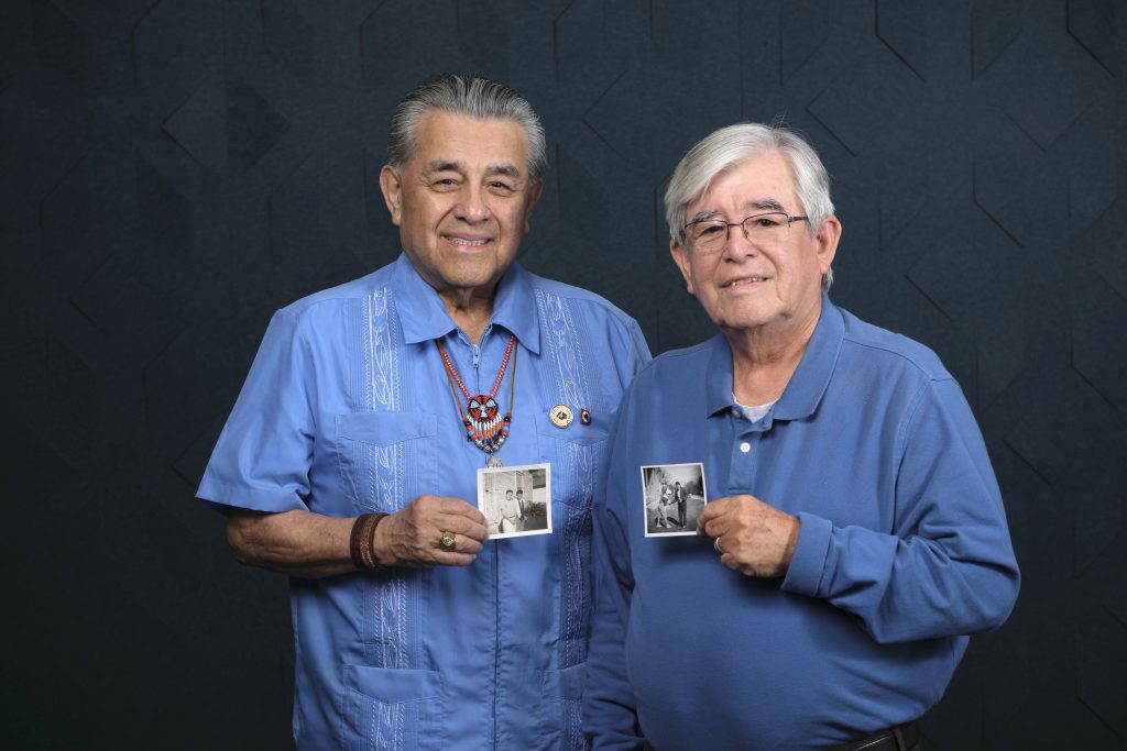 Two older men standing side-by-side smiling at the camera against a dark, textured background. The man on the left wears a light blue guayabera shirt and a beaded necklace, while the man on the right wears a blue polo shirt and glasses. Both men are holding up small, old black-and-white photographs of two younger men, presumably themselves in their youth.