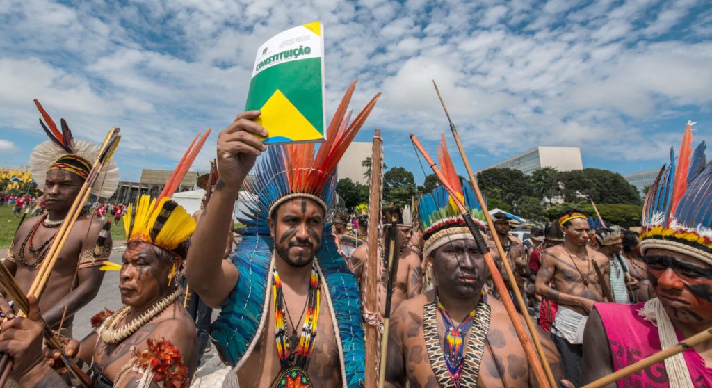 A group of Indigenous people outdoors, wearing traditional body paint, beaded jewelry, and vibrant feathered headdresses while carrying wooden bows and arrows. The man in the center is raising a booklet colored green, yellow, and blue with the word "CONSTITUIÇÃO" printed on the cover. Modern white buildings and a partly cloudy sky are visible in the background.